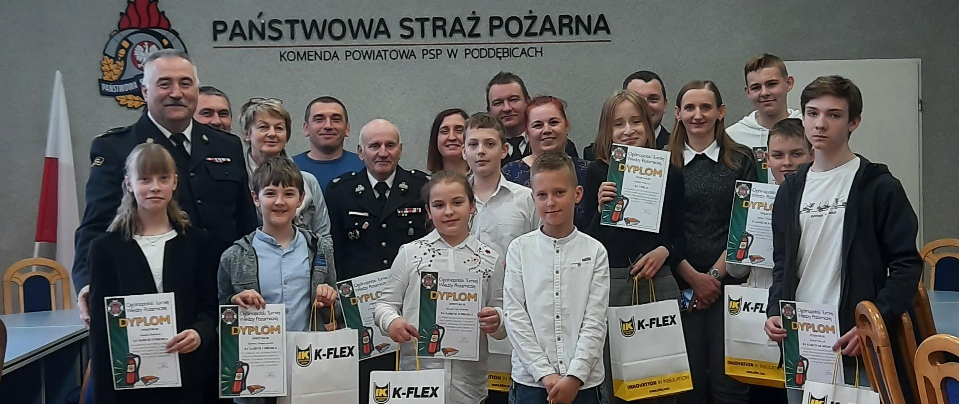 zdjęcie grupowe na sali dziewięć dzieci stoi w rękach trzymają dyplomy i torby z napisem kflex w których są nagrody. z dziećmi stoją ich opiekunowie oraz zastępca komendanta powiatowego PSP, prezes powiatowy OSP 