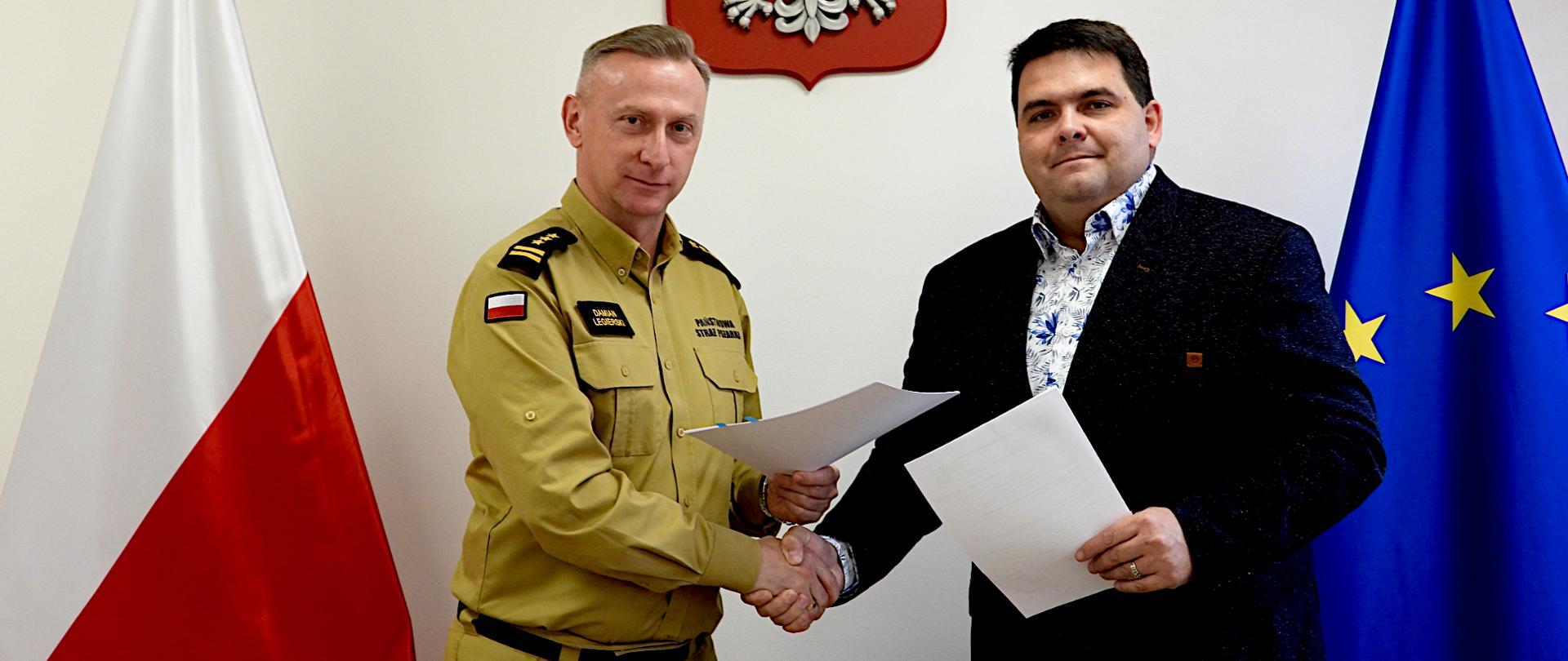 Na zdjęciu widać dwóch mężczyzn stojących obok siebie i podających sobie ręce, po podpisaniu umowy. Po lewej stronie stoi mężczyzna w mundurze w kolorze piaskowym, z naszywką polskiej flagi na ramieniu i napisem „Państwowa Straż Pożarna” na piersi jest to śląski komendant wojewódzki Państwowej Straży Pożarnej starszy brygadier Damian Legierski. Po prawej stronie stoi mężczyzna w ciemnym garniturze i koszuli we wzory - Pan Łukasz Pach Dyrektor Wojewódzkiego Pogotowia Ratunkowego w Katowicach .Obaj trzymają w dłoniach kartki papieru. W tle, za nimi, znajdują się dwie flagi – po lewej stronie flaga Polski, po prawej flaga Unii Europejskiej. 