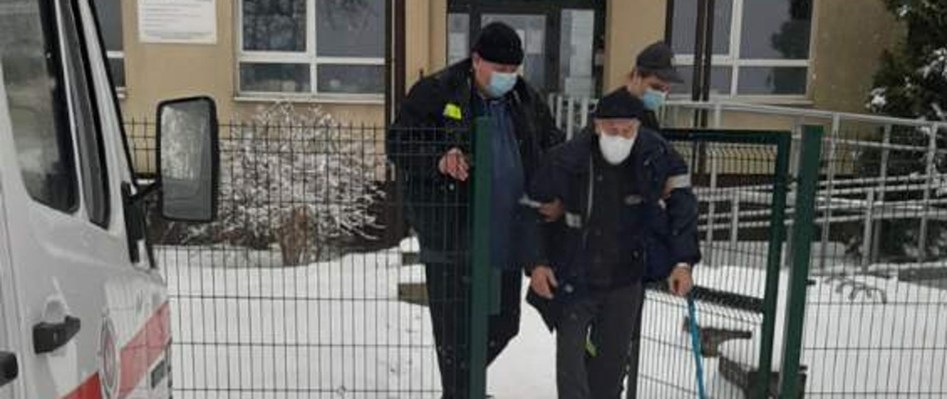 Strażak w ubraniu koszarowym prowadzi z punktu szczepień, budynku znajdującego się na drugim planie, osobę starszą. Z lewej strony fragment samochodu pożarniczego.