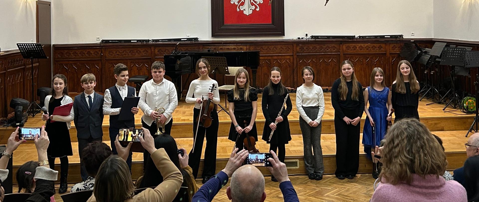 Uczniowie klasy IIIB PSM I stopnia stoją na scenie Sali Królewskiej po koncercie klasowym, trzymając instrumenty muzyczne.