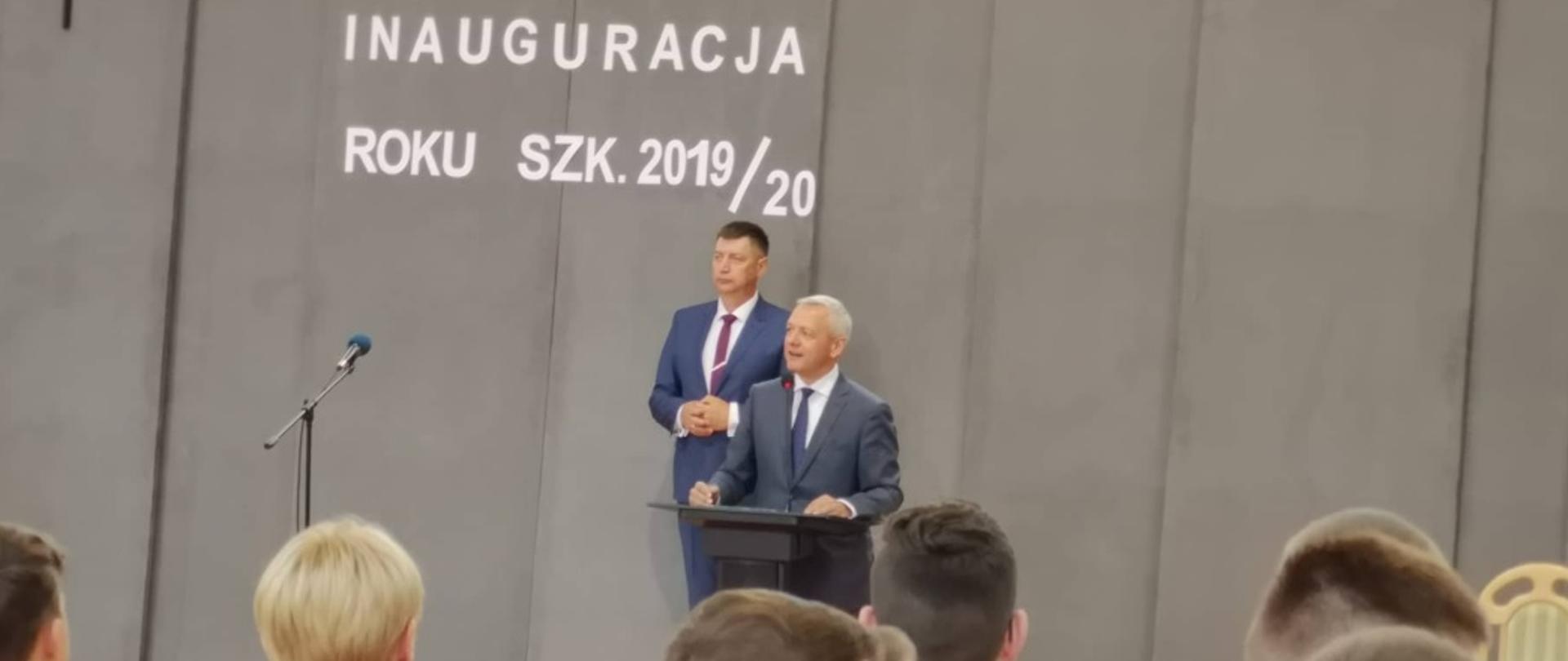 Minister Marek Zagórski na tle napisu "Inauguracja roku szkolnego 2019/20". Przed nim stoją uczniowie