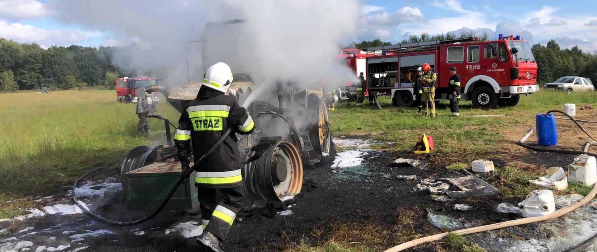 Trwa dogaszanie spalonego traktora rolniczego na polu. Strażaka z wężem polewa wodą wrak. W oddali samochody strażackie.
