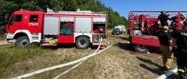 Obok akwenu wodnego na zielonej łące stoją czerwone samochody pożarnicze. Wzdłuż brzegu rozwinięte dwie linie wężowe. Z tyłu zielone drzewa. Obok przyczepy stoją strażacy.
