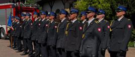 Pododdział strażaków stoi w dwuszeregu. Na dalszym planie wóz strażacki i budynek.
