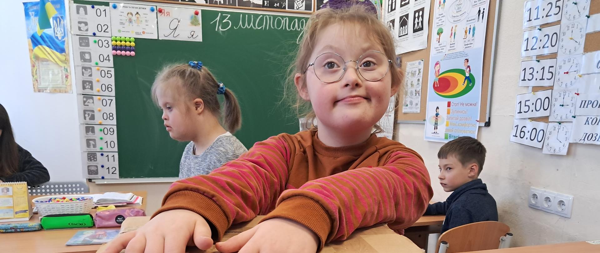 W klasie w ławkach siedzą dzieci z niepełnosprawnością; na pierwszym planie dziewczynka trzyma ręce na paczce z pomocą materialną