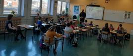 Na zdjęciu widać klasę szkolną na zajęciach z edukacji dla bezpieczeństwa. Na końcu sali znajduje się strażak w umundurowaniu, który prowadzi zajęcia edukacyjne. 