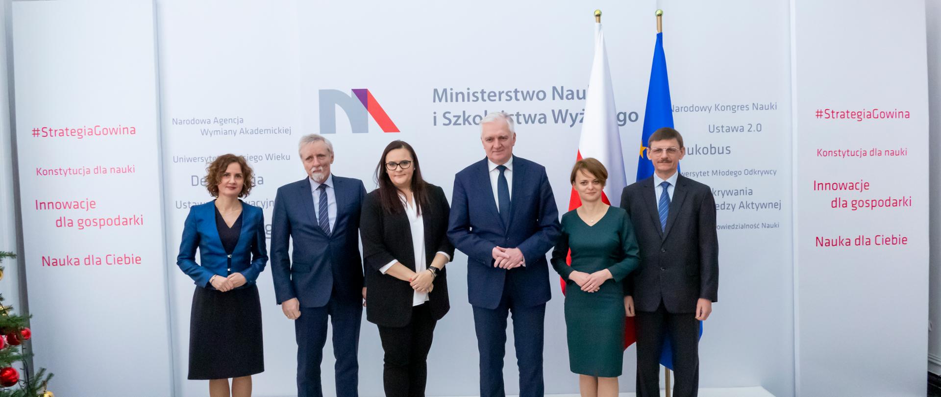 Zdjęcie grupowe. 6 osób wśród nich między innymi minister Małgorzata Jarosińska-Jedynak, wicepremier Gowin, minister Emilewicz. Osoby stoją na scenie, za nimi stoi flaga Polski i Unii Europejskiej oraz ścianka z napisem Ministerstwo Nauki i Szkolnictwa Wyższego. 