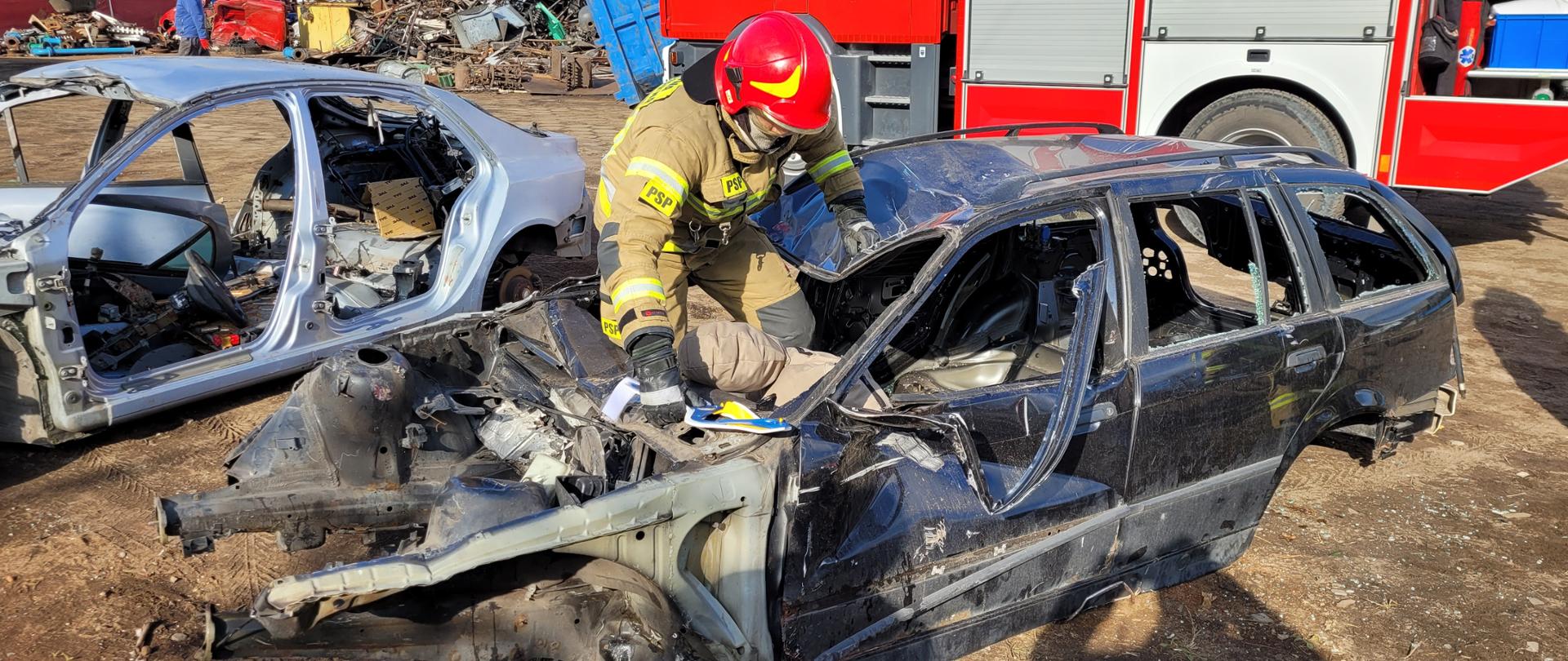 Na pierwszym planie zdjęcia widoczny jest strażak- ratownik w ubraniu specjalnym oraz czerwonym hełmie. Strażak dostaje się do wraku pojazdu przez otwór po przedniej szybie auta. W prawym ręku trzyma kołnierz ortopedyczny dla poszkodowanego, w środku pojazdu znajduje się manekin imitujący osobę poszkodowaną. W tle widać czerwony wóz strażacki z otwartą jedną skrytką sprzętową. Po lewej stronie zdjęcia widać składowisko złomu.