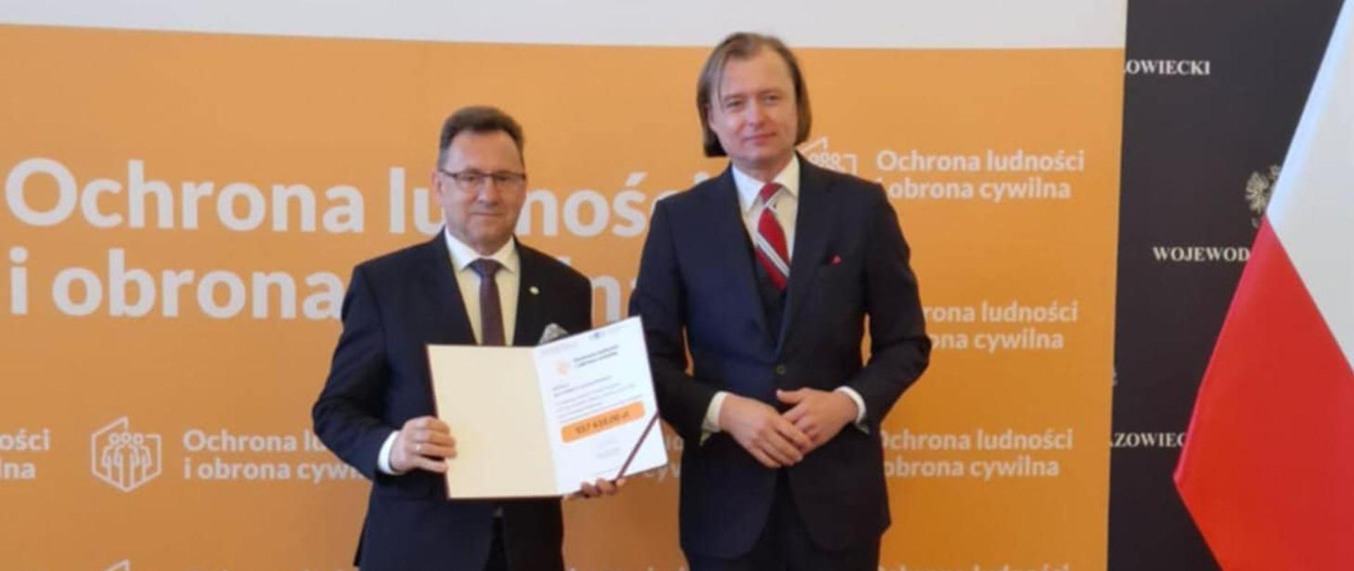 Na zdjęciu widzimy dwóch mężczyzn w strojach formalnych stojących obok siebie na tle dużej, pomarańczowo-białej ściany z napisami w języku polskim.
Mężczyzna po lewej stronie ma okulary i jest ubrany w ciemny garnitur z białą koszulą i ciemnym krawatem w drobny wzór. Trzyma w rękach otwarty folder z informacją o kwocie dotacji, który jest skierowany w stronę drugiego mężczyzny.
Mężczyzna po prawej stronie ma jasne, dłuższe włosy zaczesane do tyłu. Jest ubrany w ciemny garnitur z białą koszulą i czerwonym krawatem z ciemnym wzorem oraz ma przypiętą małą odznakę na klapie. Patrzy na mężczyznę po lewej.
W tle, po prawej stronie, widać również fragment flagi Polski (czerwień i biel). Na pomarańczowej ścianie dominują napisy związane z "Ochrona ludności i obrona cywilna". Całe ujęcie sugeruje, że jest to moment publiczny, związany z podpisaniem dofinansowania.