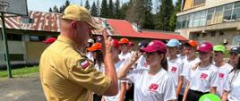Zdjęcie zrobione na zewnątrz, na boisku sportowym, podczas uroczystej zbiórki uczestników obozu Młodzieżowych Drużyn Pożarniczych. W szeregu stoją uczestnicy obozu szkoleniowo-wypoczynkowego młodzieżowych drużyn pożarniczych. Oficer Państwowej Straży Pożarnej salutuje dziewczynce, która jest uczestnikiem obozu MDP.