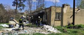 Widoczny budynek murowany, w koło dużo śmieci i gruzu, strażacy pracujący w zadymieniu w sprzęcie ochronnym izolacyjnym