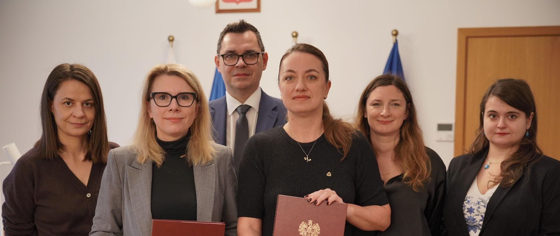 Na zdjęciu widzimy grupę osób stojących w biurze, w tle z polską flagą i flagą Unii Europejskiej. Na pierwszym planie, wśród grupy, znajduje się Pani Monika Sikora, wiceministera funduszy i polityki regionalnej, która trzyma w ręku dokument z godłem Polski.Obok stoją inne osoby w eleganckich strojach. Zdjęcie ma charakter formalny i może pochodzić z wydarzenia związanego z wprowadzeniem dostępności architektonicznej zabytków.