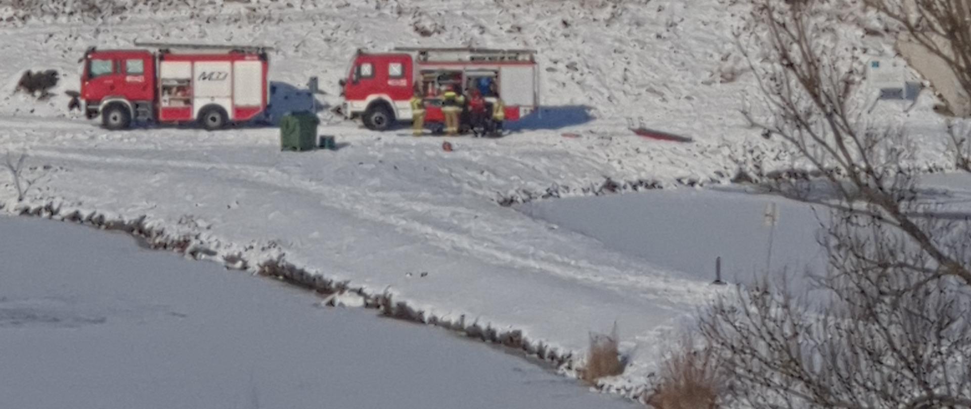 Dwa samochody gaśnicze Państwowej Straży Pożarnej stojące pomiędzy 2 zbiornikami wodnymi. Funkcjonariusze stoją przed samochodem pożarniczym. Przed i za zbiornikiem wodnym znajdują się drzewa. 