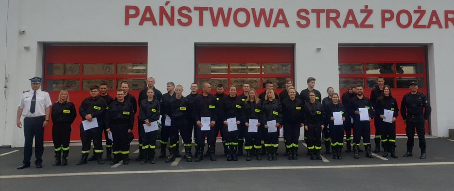 Zdjęcie przedstawia uczestników szkolenia podstawowego strażaków ratowników OSP wraz z Komendantem Powiatowym PSP w Obornikach oraz Dowódcą JRG Oborniki na tle budynku strażnicy. 