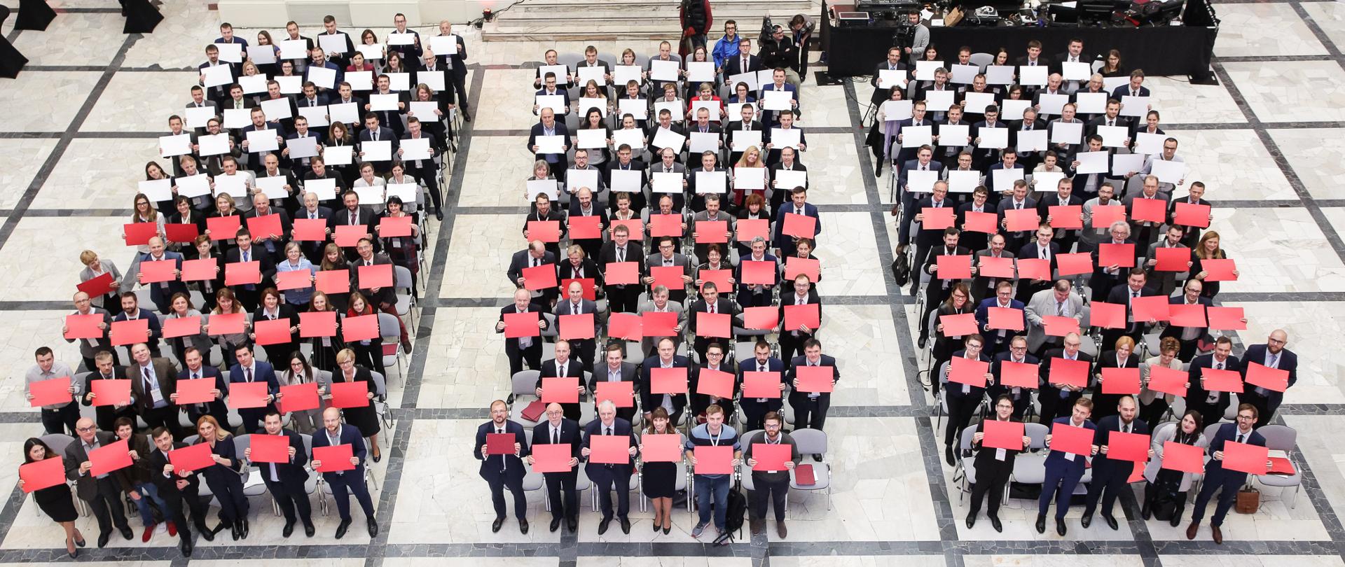 Na zdjęciu trzy rzędy stojących ludzi - każdy z nich trzyma biały lub czerwony karton. Dzięki temu tworzy się biało-czerwona flaga.