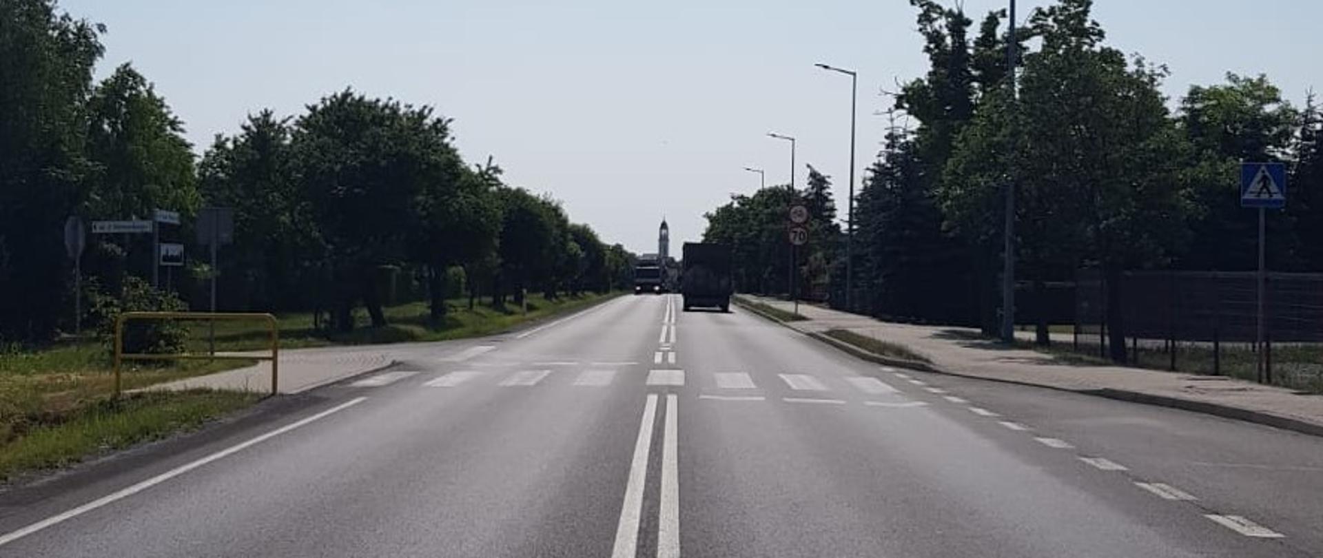 Zdjęcie wykonane z poziomu ziemi. Na środku jezdni widoczne białe linie, w oddali widoczne przejście dla pieszych, z prawej strony chodnik oraz dwa pojazdy ciężarowe.