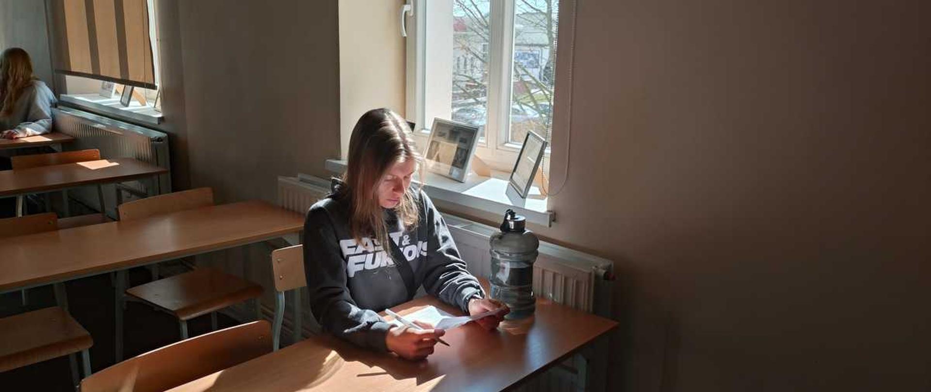 Zdjęcie przedstawia wnętrze jasnej sali lekcyjnej lub wykładowej w świetle dziennym. W centrum uwagi znajduje się młoda kobieta siedząca przy szkolnej ławce, która w skupieniu czyta kartkę papieru.
Szczegóły kompozycji
Postać: W centralnej części siedzi dziewczyna o długich, jasnobrązowych włosach. Ma na sobie ciemnoszarą bluzę z kapturem z białym napisem „FAST & FURIOUS”. W dłoniach trzyma białą kartkę i długopis, sprawiając wrażenie osoby piszącej test lub analizującej notatki.
Otoczenie:
Dziewczyna siedzi przy klasycznej, drewnianej ławce o jasnym blacie. Obok niej stoi duża, ciemna butelka na wodę (typu bidon).
W tle, po lewej stronie, widać fragment innej osoby siedzącej w dalszej ławce oraz rzędy pustych stolików i krzeseł.
Po prawej stronie znajduje się duże okno, przez które wpada intensywne, naturalne światło słoneczne, tworząc wyraźne cienie na blacie i podłodze. Na parapecie stoją oprawione zdjęcia lub dyplomy.
Oświetlenie i kolorystyka: Zdjęcie charakteryzuje się dużym kontrastem. Silne światło słoneczne ociepla barwy drewna i mocno oświetla postać dziewczyny, podczas gdy reszta sali pozostaje w delikatnym półcieniu.
Nastrój
Zdjęcie oddaje atmosferę ciszy, spokoju i nauki. Surowość sali lekcyjnej jest przełamana przez ciepłe, wpadające przez okno promienie słońca.