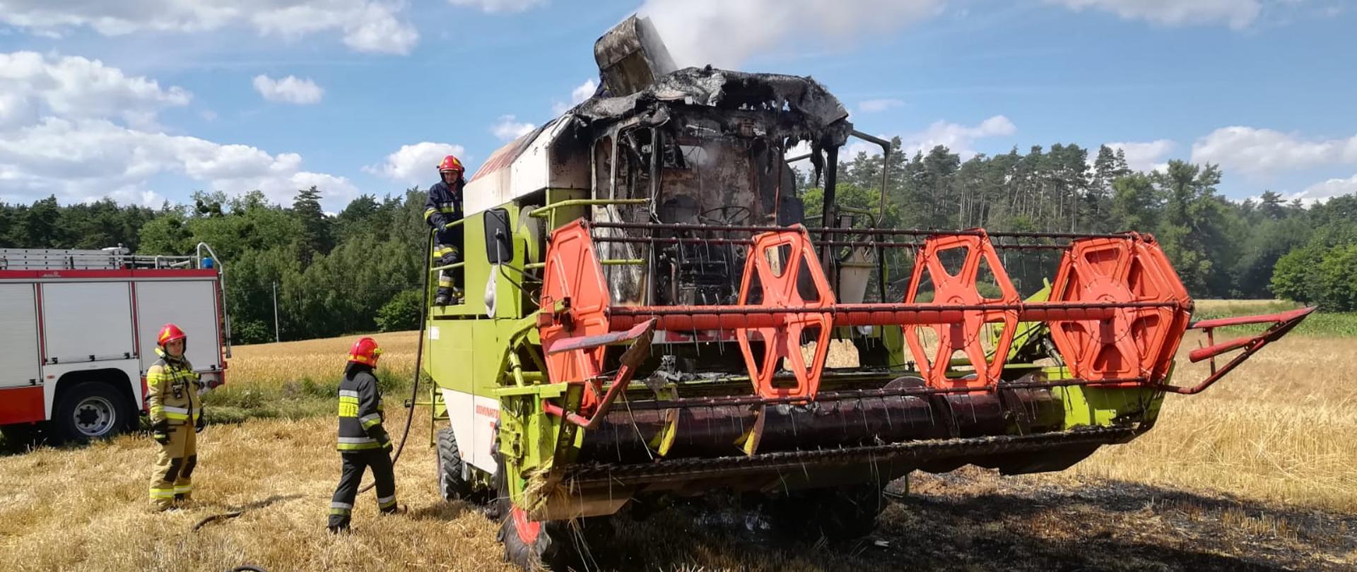 Zielony częściowo spalony kombajn do zboża, który stoi na polu. Obok niego stoi czerwony samochód strażacki a trzech strażaków prowadzi działania gaśnicze.