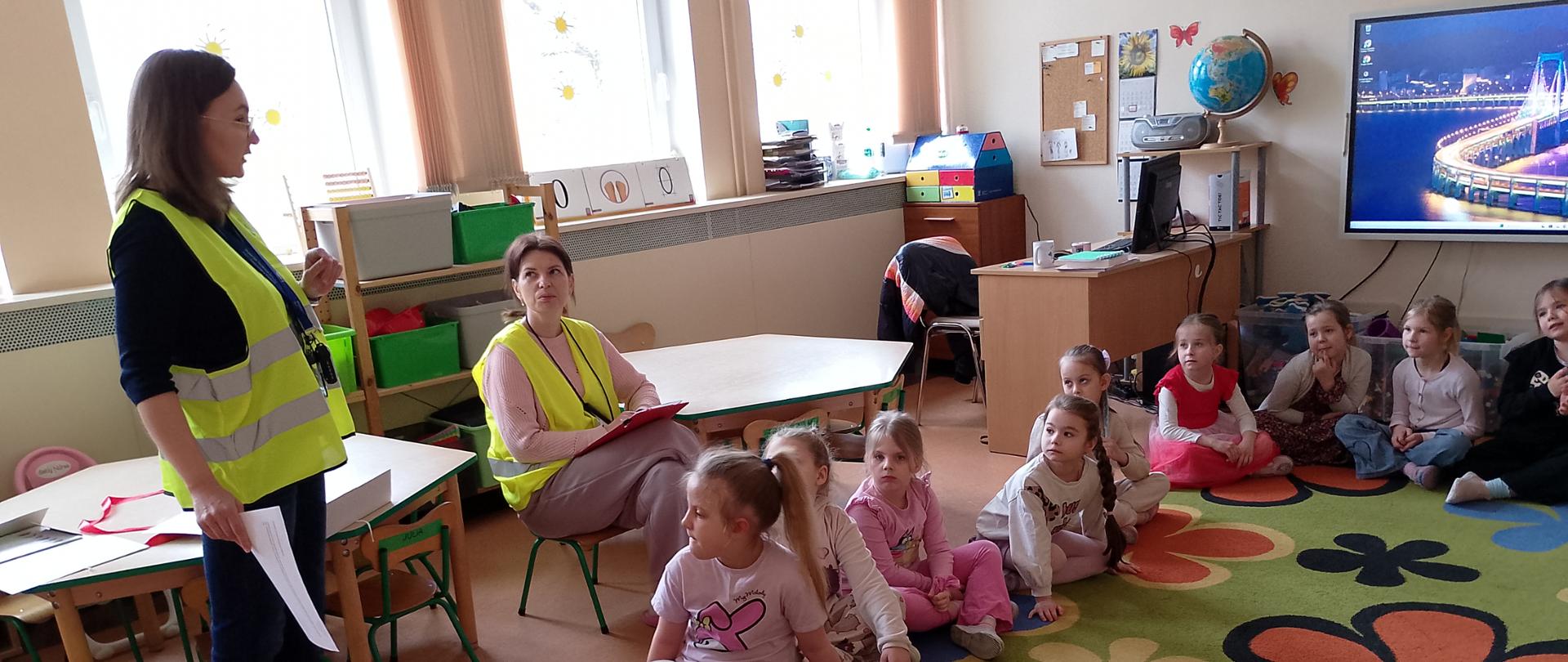 Zdjęcie przedstawia salę przedszkolną. Po lewej stronie zdjęcia znajduje się dwóch pracowników WIORiN. Po prawej stronie zdjęcia znajdują się dzieci siedzące na dywanie. Dzieci słuchają pracownika WIORiN.