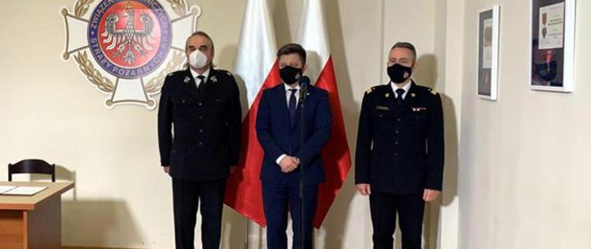 Trzech mężczyzn stoi w rzędzie na tle dwóch flag Rzeczypospolitej Polski i logo ZOSP RP. Mężczyzna stojący w środku ubrany jest w granatowy garnitur , mężczyźni stojący po jego bokach są ubrani w mundury wyjściowe. Wszyscy mają na twarzach maseczki zasłaniające twarz i usta.