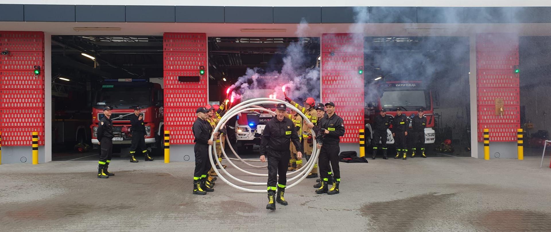 Strażacy stoją na przeciwko siebie i trzymają w rękach zwinięty w spiralę waż. W środku stoi wyróżniony strażak. W tle budynek remizy strażackiej z widocznymi pojazdami pożarniczymi.