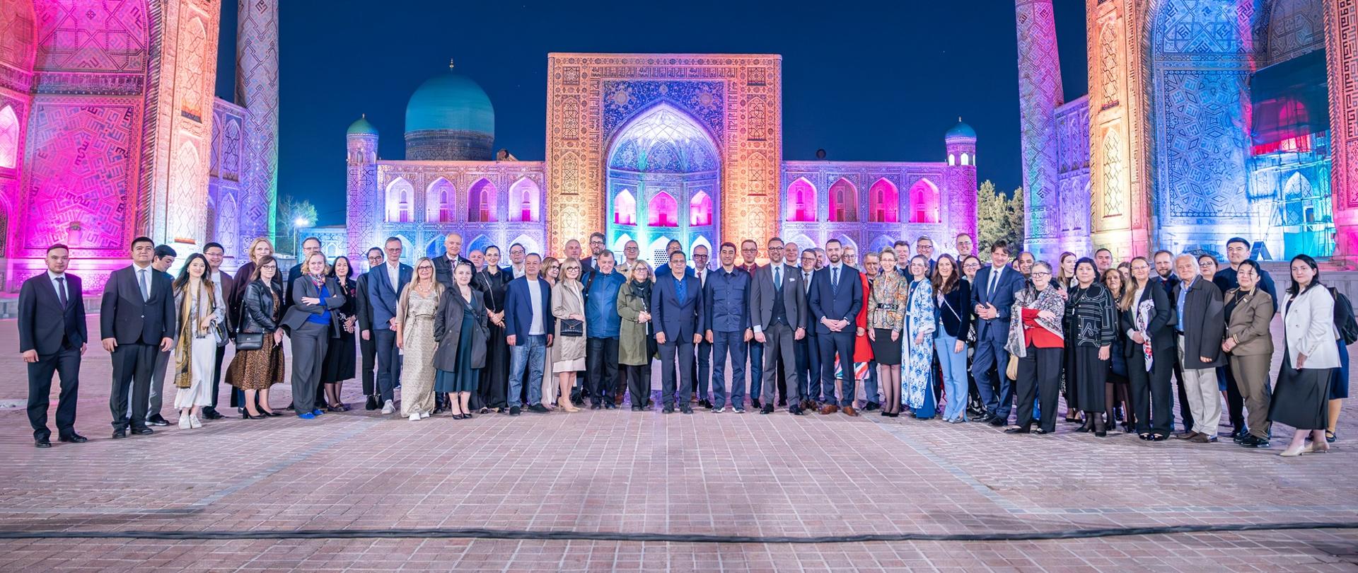 Na dworze, na placu wyłożonym czerwono-brązową kostką stoi grupa kilkudziesięciu osób, za nimi bardzo kolorowe budynki we wschodnim stylu.