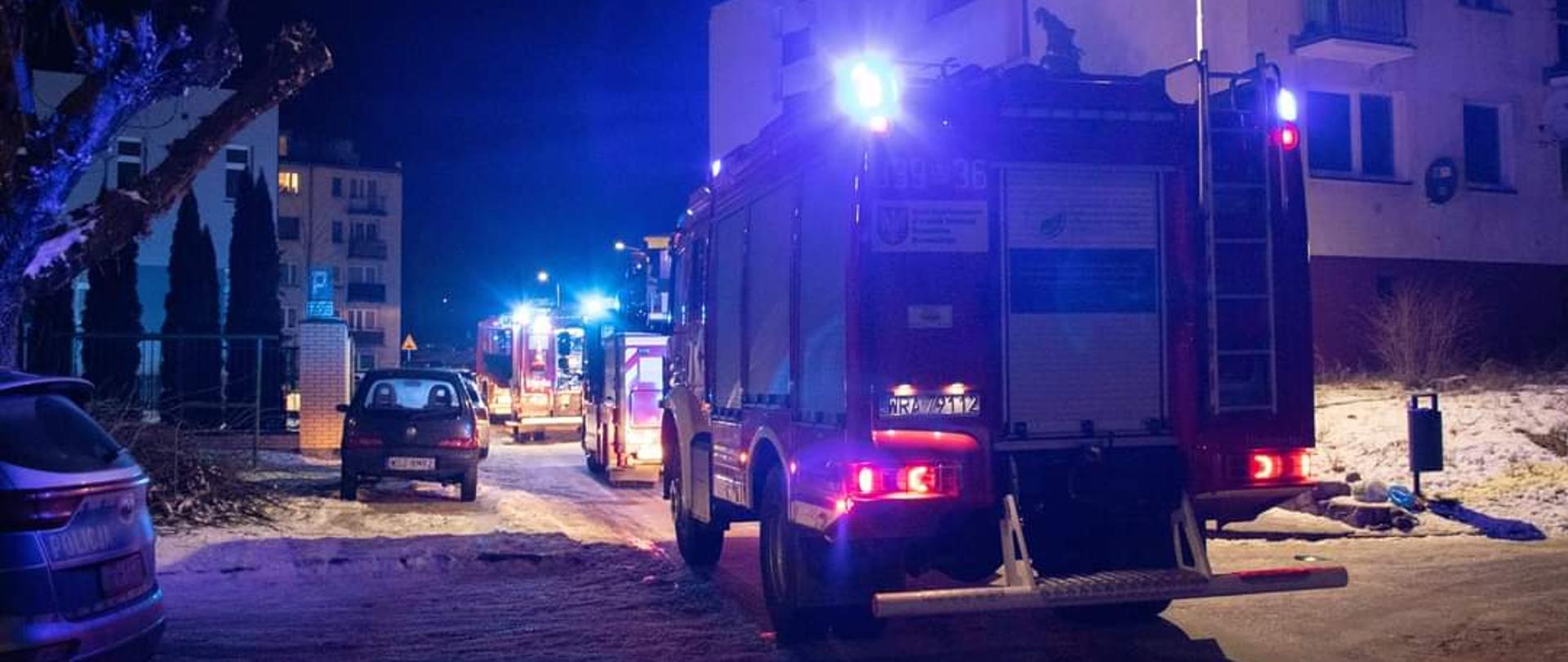 Zdjęcie wykonane w porze nocnej w miejscowości Pionki. Na zdjęciu widać jedno z osiedli bloków mieszkalnych w Pionkach przy ulicy Kozienickiej. Przy blokach widać zaparkowane pojazdy ratowniczo - gaśnicze Państwowej i Ochotniczej Straży Pożarnej. Pojazdy w kolorze czerwonym z białymi i srebrnymi elementami zabudowy. Pojazdy zapalone mają również niebieskie światła alarmowe. Obok pojazdów ratowniczych przy drodze zaparkowane są pojazdy należące do mieszkańców osiedla. Po lewej stronie zdjęcia widać również tył radiowozu Policji. 