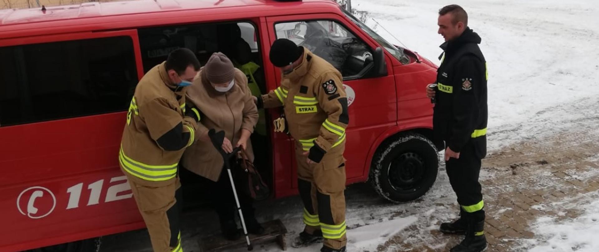 Obraz przestawia strażaków pomagających osobie starszej wysiąść z samochodu pożarniczego.
