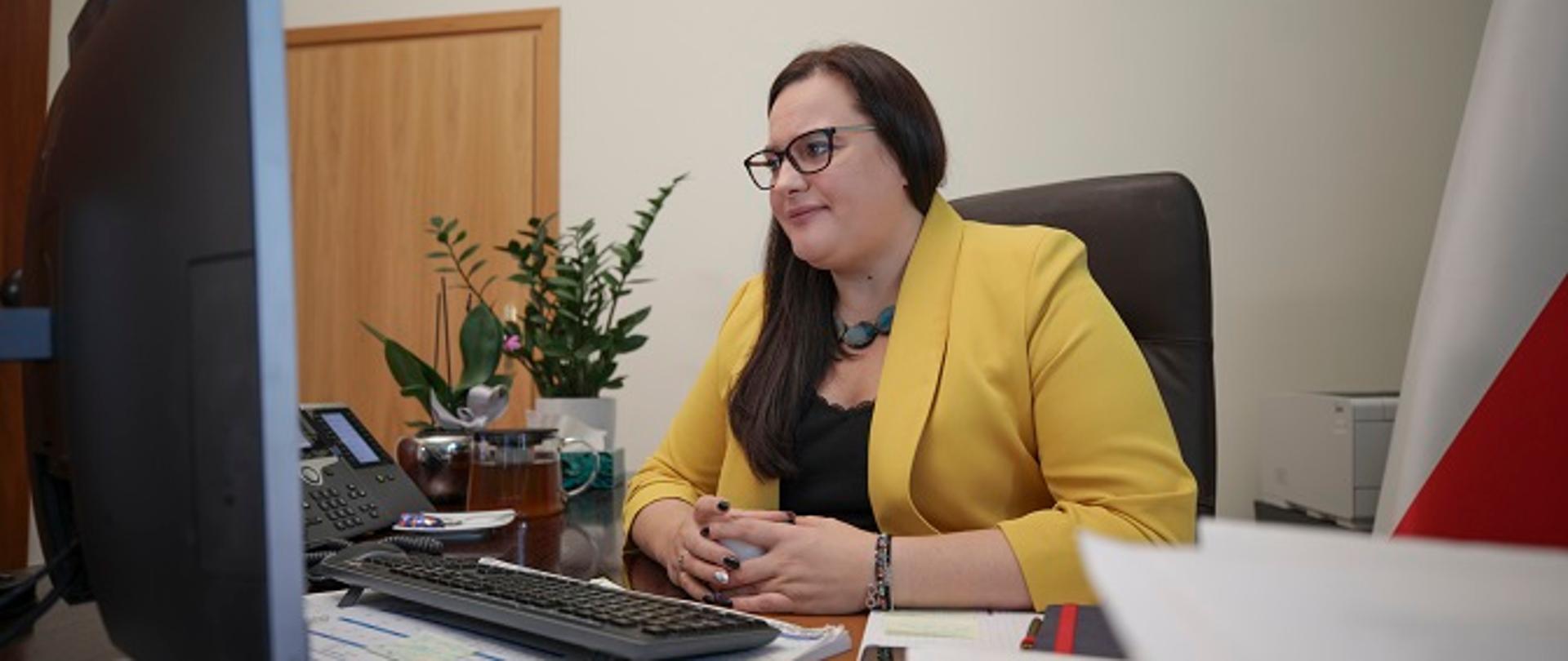 Minister Małgorzata Jarosińska-Jedynak siedzi przy swoim biurku, przed komputerem, gdzie online odbywa się wideokonferencja z podmiotami, które przystąpiły do Partnerstwa na rzecz Dostępności.