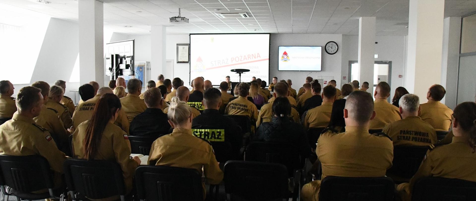 Strażacy w mundurach koloru musztardowego oraz czarnego siedzą na krzesłach obok siebie na ekranie multimedialnym wyświetlany jest slajd.