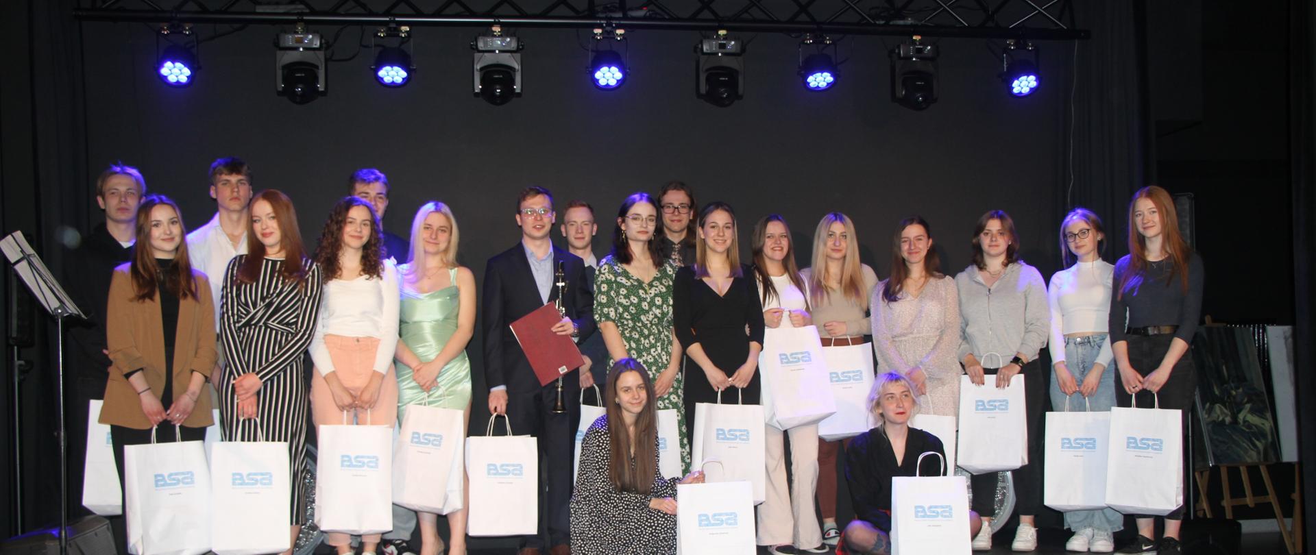 na zdjeciu kilkunastoosobowa grupa dziewcząt i chłopców stoi na scenie, w rękach trzymają białe torby z niebieskim napisem BSA