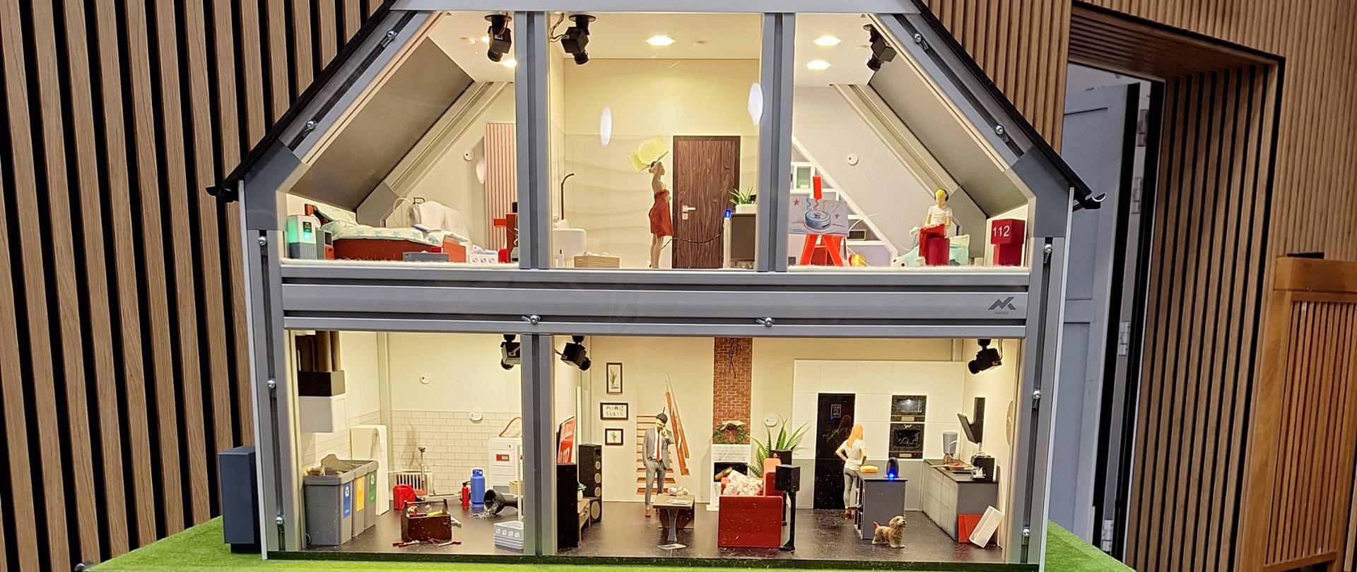 Na zdjęciu widać makietę domu jednorodzinnego w przekroju, ustawioną na ekspozycyjnym postumencie.
Makieta przedstawia dwukondygnacyjny dom z poddaszem, pokazany „od przodu” bez ściany, dzięki czemu widać szczegółowo wnętrza pomieszczeń. W środku znajdują się miniaturowe meble, sprzęty domowe oraz figurki ludzi, które imitują codzienne czynności (np. gotowanie, odpoczynek, prace domowe). Pomieszczenia są oświetlone, co nadaje makiecie realistyczny charakter. Całość stoi na zielonej podstawie imitującej trawnik.
Pod makietą znajduje się drewniany cokół z tablicą informacyjną. Widnieje na niej logo oraz napis:
Wojewódzki Fundusz Ochrony Środowiska i Gospodarki Wodnej w Warszawie
oraz informacja, że jest to „zakup symulatora zagrożeń pożarowych i edukacji ekologicznej”, dofinansowany przez WFOŚiGW w Warszawie.
Całość sugeruje, że makieta służy celom edukacyjnym, prawdopodobnie do szkoleń lub pokazów związanych z bezpieczeństwem pożarowym i ekologią w gospodarstwach domowych.
