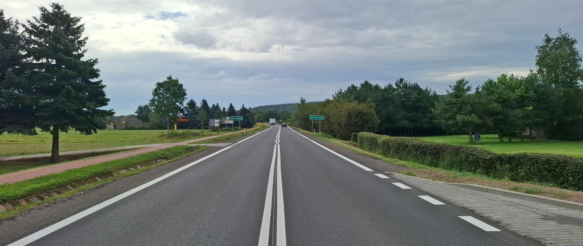 DK9 Nietulisko - prosta jednojezdniowa droga asfaltowa. Wzdłuż drogi namalowane białe linie oznakowania poziomego - ciągle i przerywane. W oddali samochody, Po obu stronach drogi drzewa i zieleń.
