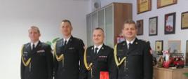 Na zdjęciu widać czterech strażaków w umundurowaniu galowym. Strażacy znajdują się w biurze. Umundurowanie jest koloru czarnego ze złotymi sznurami na klatce piersiowej. Jeden ze strażaków trzyma czerwoną aktówkę. 