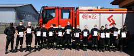 Na zdjęciu widać strażaków ochotników trzymających w rękach dyplomy ukończenia kursu dowódców OSP. W tle stoi samochód strażacki.