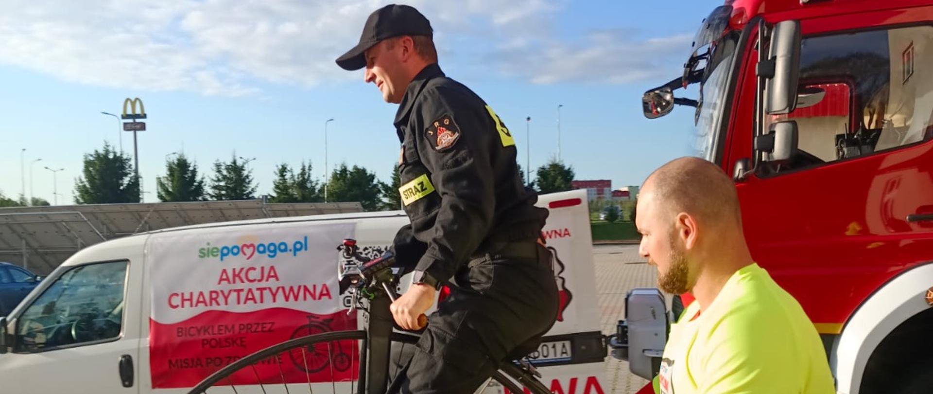 Na zdjęciu strażak w umundurowaniu koszarowym, który jedzie na starym rowerze z wielkim przednim kołem, tzw. bicyklu. Obok idzie mężczyzna w jaskrawej koszulce i różowych spodenkach, który asekuracyjnie trzyma rower. W tle stoi czerwony wóz strażacki oraz biały samochód dostawczy z banerem „Akcja charytatywna” i logo „siepomaga.pl”. Nad całą sceną niebo jest częściowo zachmurzone, ale jasne i pogodne.