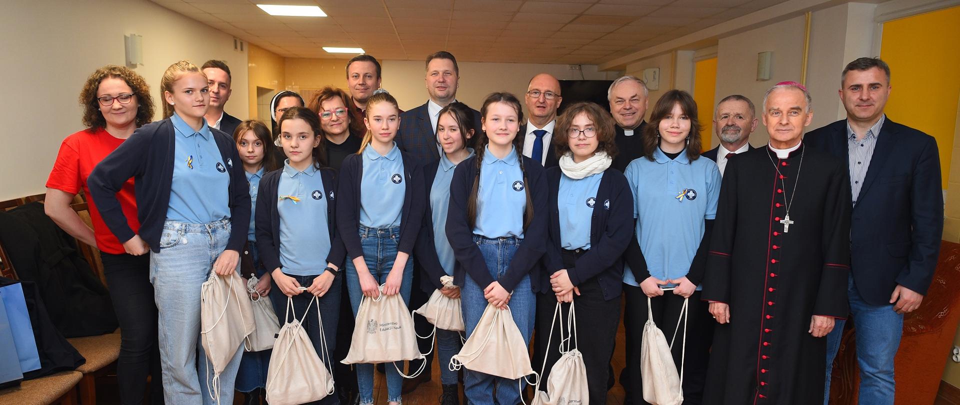 Na zdjęciu dzieci w towarzystwie księdza i ministra stoją w korytarzu.