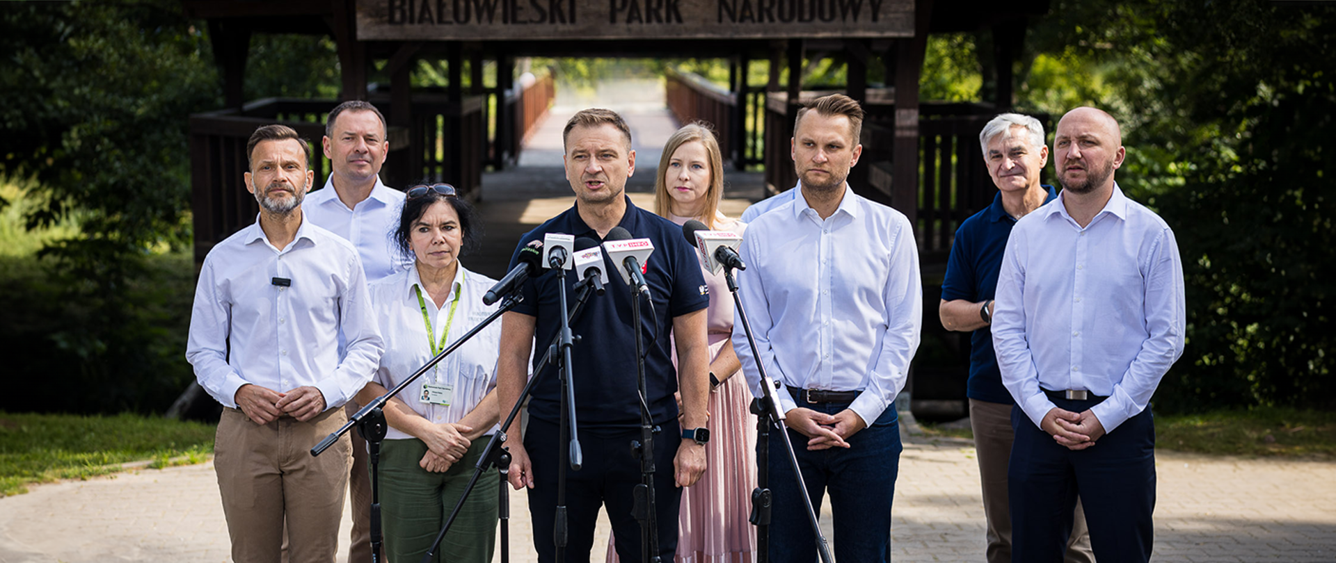 Uczestnicy konferencji prasowej 3 lipca 2024 w Białowieży stoją przed mikrofonami. Na środku Minister Sportu i Turystyki Sławomir Nitras. Za nimi wejście do Białowieskiego Parku Narodowego. Widoczny mostek a nad nim napis "Białowieski Park Narodowy".