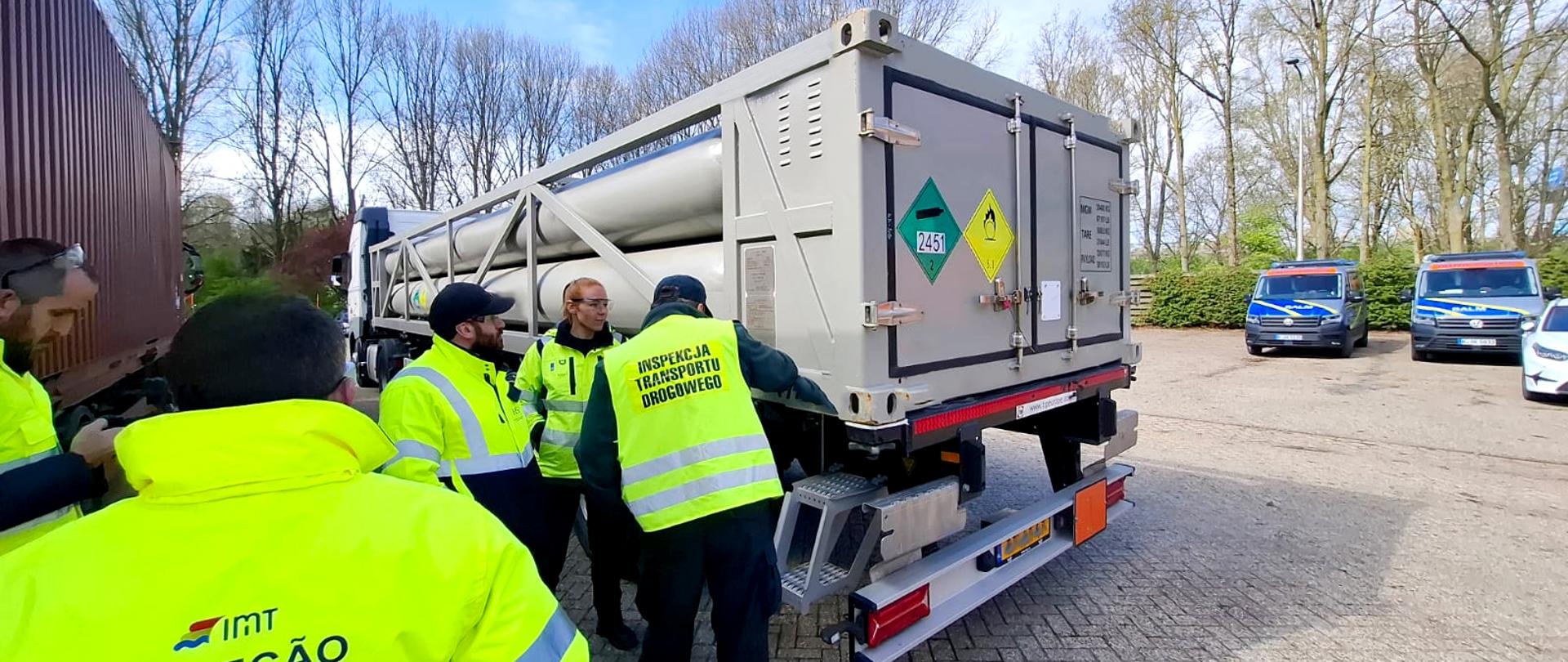 Inspektorzy sprawdzają tył pojazdu z kontenerem-cysterną oznakowanym tablicami ADR, oceniając stan techniczny i oznaczenia.