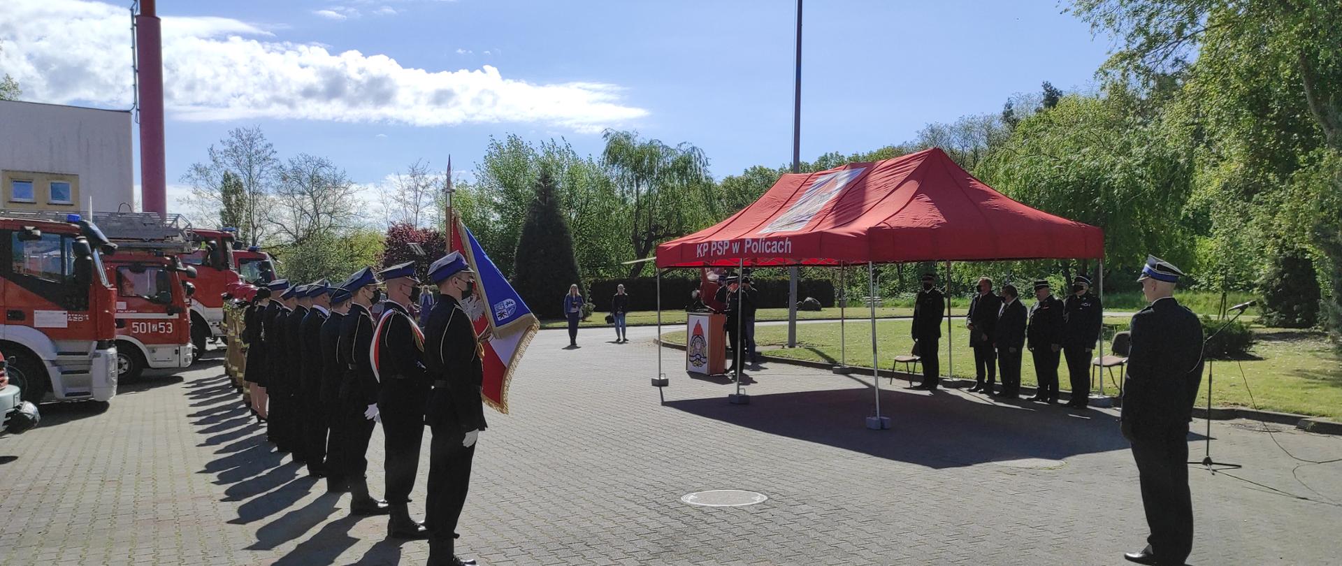 Na zdjęciu widoczny plac z polbruku i fragment budynku strażnicy. Przed garażami budynku stojące strażackie pojazdy, przed nimi widoczne plecy stojących w równym szeregu, wyprostowanych strażaków, część z nich w ciemnogranatowym umundurowaniu galowym z żółtymi sznurami i rogatywkami na głowach, część stojących w głębi w piaskowym ubraniu bojowym w czerwonych hełmach na głowie. Na pierwszym planie po lewej stronie sztandar na drzewcu, po prawej stojącego bokiem strażaka w mundurze galowym ze sznurem i rogatywce. Przed nim czerwone zadaszenie otwartego namiotu z napisem KP PSP w Policach, pod którym widać stojące postaci, część z nich w mundurach. Dalej widoczna mównica z logo Państwowej Straży Pożarnej i przemawiający Komendant Powiatowy PSP w Policach st. kpt. Piotr Maciejczyk. W głębi zdjęcia zieleń drzew i krzewów, niebo błękitne z nielicznymi białymi obłokami.