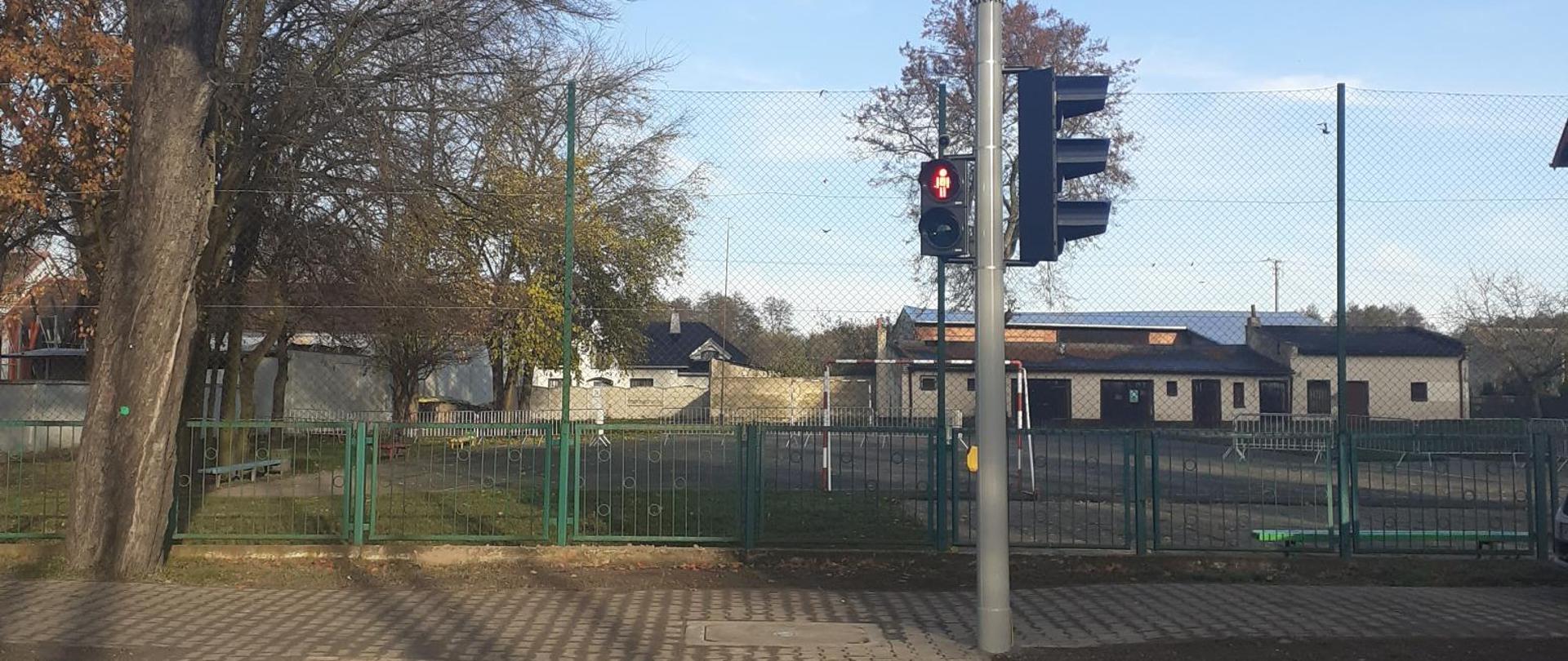 zdjęcie przedstawia przejście dla pieszych z sygnalizacją świetlną. Światło czerwone. W tle widoczne zabudowania.