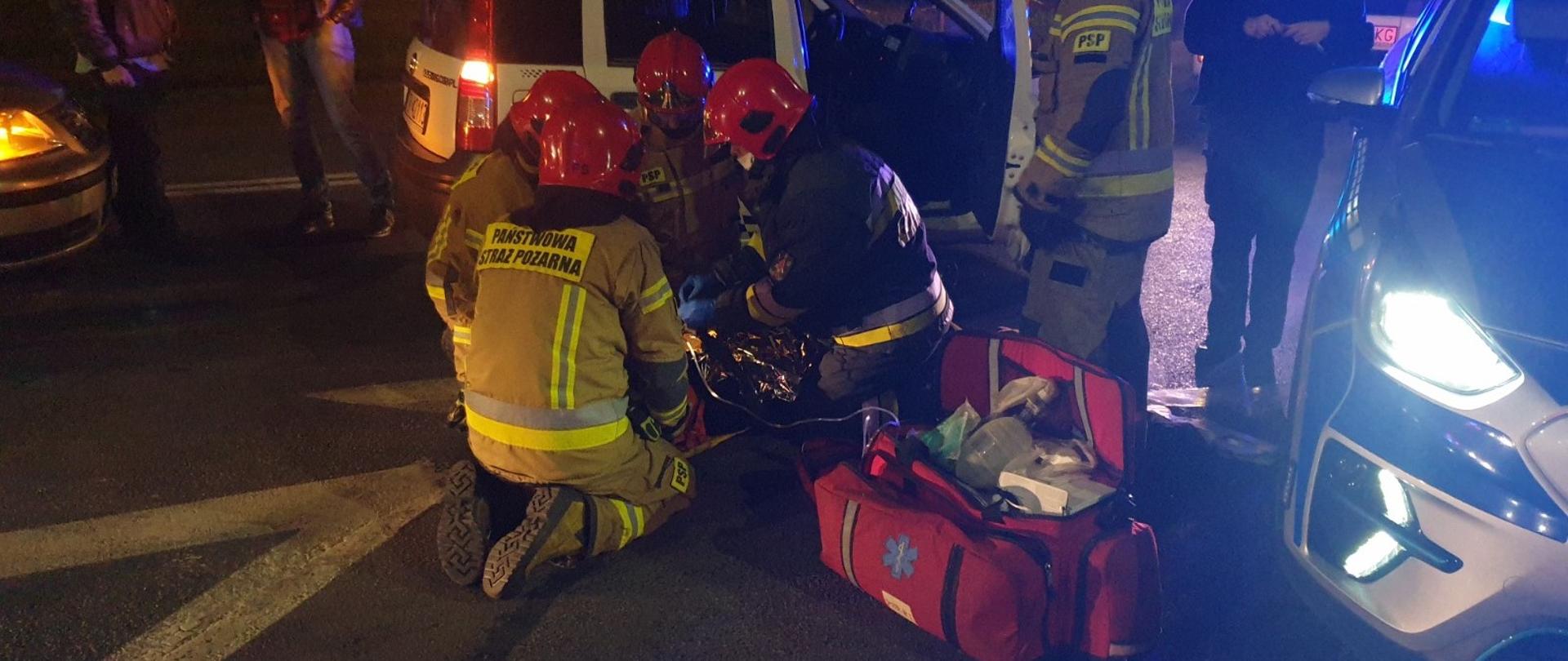 Zdjęcie obrazuje czterech strażaków ubranych w ubrania bojowe, rękawiczki lateksowe i maseczki jednorazowe, udzielających kwalifikowanej pierwszej pomocy osobie poszkodowanej w wypadku samochodowym. Obok nich leży czerwona torba medyczna. A za nimi jest funkcjonariusz policji. Wszyscy są otoczeni samochodami osobowymi w tym radiowozem policji. 