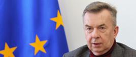 Minister Wieczorek siedzi za stołem, za nim przed ścianą flaga UE.