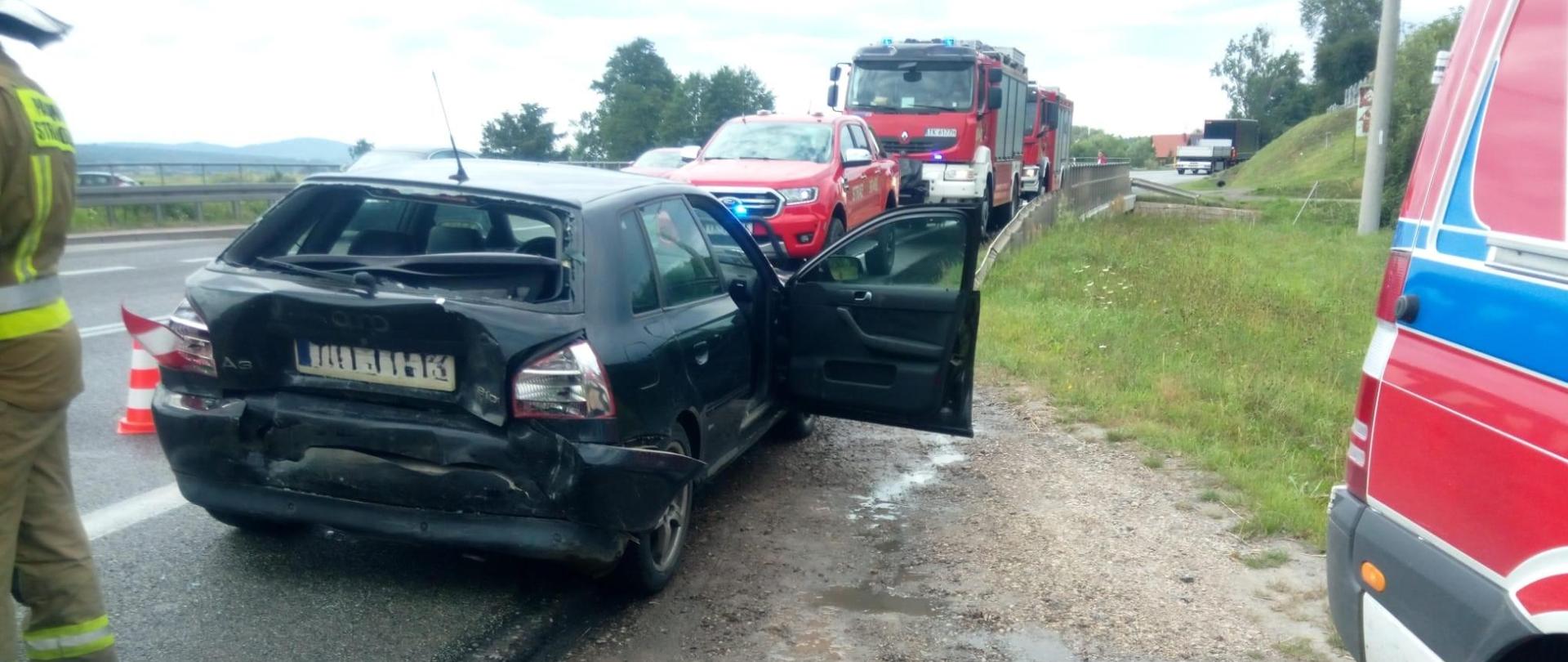 Zdjęcie przedstawia drogę krajową i ustawione na niej samochody. Kolejno stoją: uszkodzony samochód osobowy biorący udział w zderzeniu i trzy samochody pożarnicze. Po prawej stronie widać fragment ambulansu.