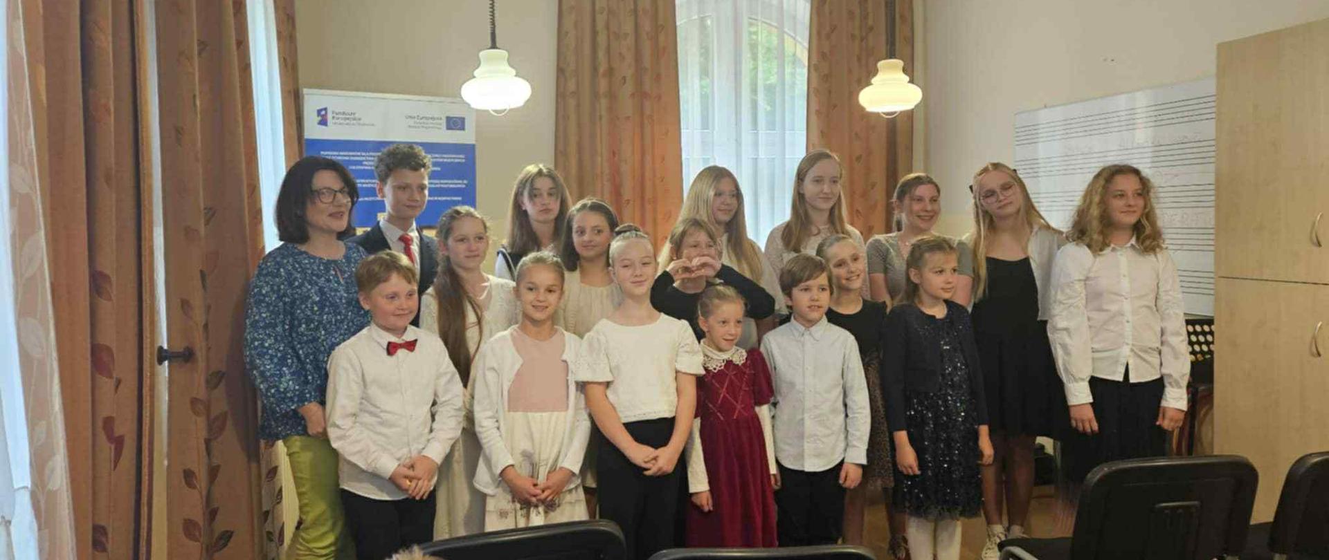 Zdjęcie przedstawia grupę dzieci i młodzieży oraz jedną dorosłą kobietę, stojących w jasnym pomieszczeniu, prawdopodobnie sali lekcyjnej lub prób. Po lewej stronie kadru stoi uśmiechnięta kobieta w okularach, ubrana w kwiecistą bluzkę i jasne spodnie. Obok niej, w kilku rzędach, ustawionych jest około piętnastu młodych osób. Są to zarówno chłopcy, jak i dziewczęta, w różnym wieku. Większość z nich jest elegancko ubrana, co sugeruje formalną okazję – dziewczynki mają sukienki lub spódniczki, a chłopcy koszule, niektórzy z muszkami. Patrzą w stronę obiektywu, a na ich twarzach widać uśmiechy lub spokojne wyrazy.
Tło pomieszczenia jest jasne. Po lewej stronie, za kobietą, widoczny jest fragment plakatu lub tablicy informacyjnej z logo. Ściany są kremowe lub jasnobrązowe. W tle znajdują się okna zasłonięte beżowymi zasłonami, a także meble, w tym szafa lub regał. Na ścianie po prawej stronie wisi duża tablica z pięciolinią. Z sufitu zwisają dwie lampy Na pierwszym planie, w dolnej części zdjęcia, widać fragmenty ciemnych krzeseł, co sugeruje, że zdjęcie zostało wykonane z perspektywy siedzącej osoby. Całość kadru jest dobrze oświetlona, a atmosfera wydaje się być uroczysta i pozytywna.