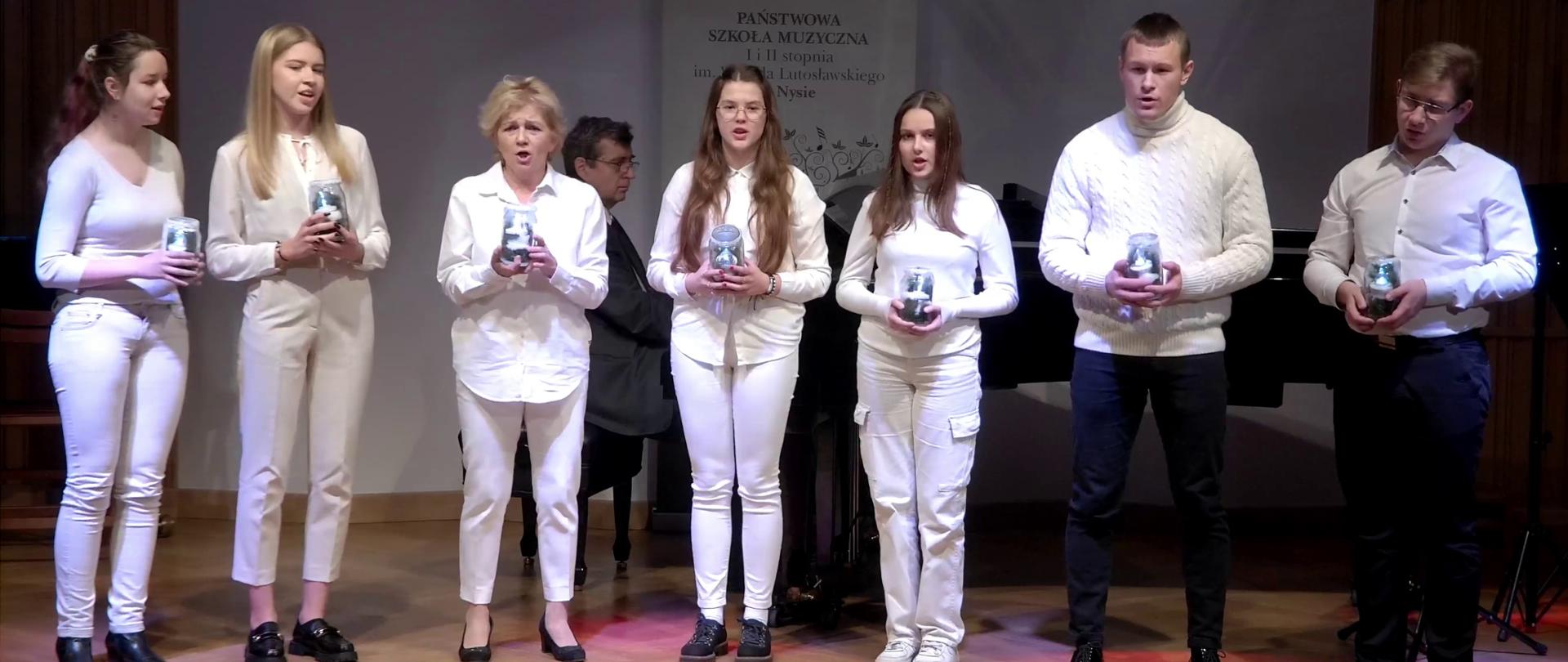 Zdjęcie - Koncert Świąteczny. Na pierwszym planie zespół wokalny. Od lewej pięć kobiet ubranych na biało oraz dwaj panowie. Wszyscy trzymają w rękach lampiony i śpiewają. Z tyłu pianista - akompaniator gra na czarnym fortepianie. W tle baner PSM Nysa.