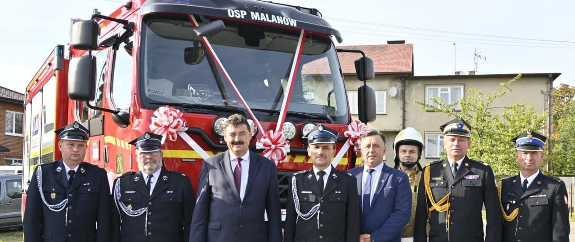 Przed nowym wozem strażackim w rzędzie do zdjęcia grupowego pozują strażacy ochotnicy oraz urzędnicy.