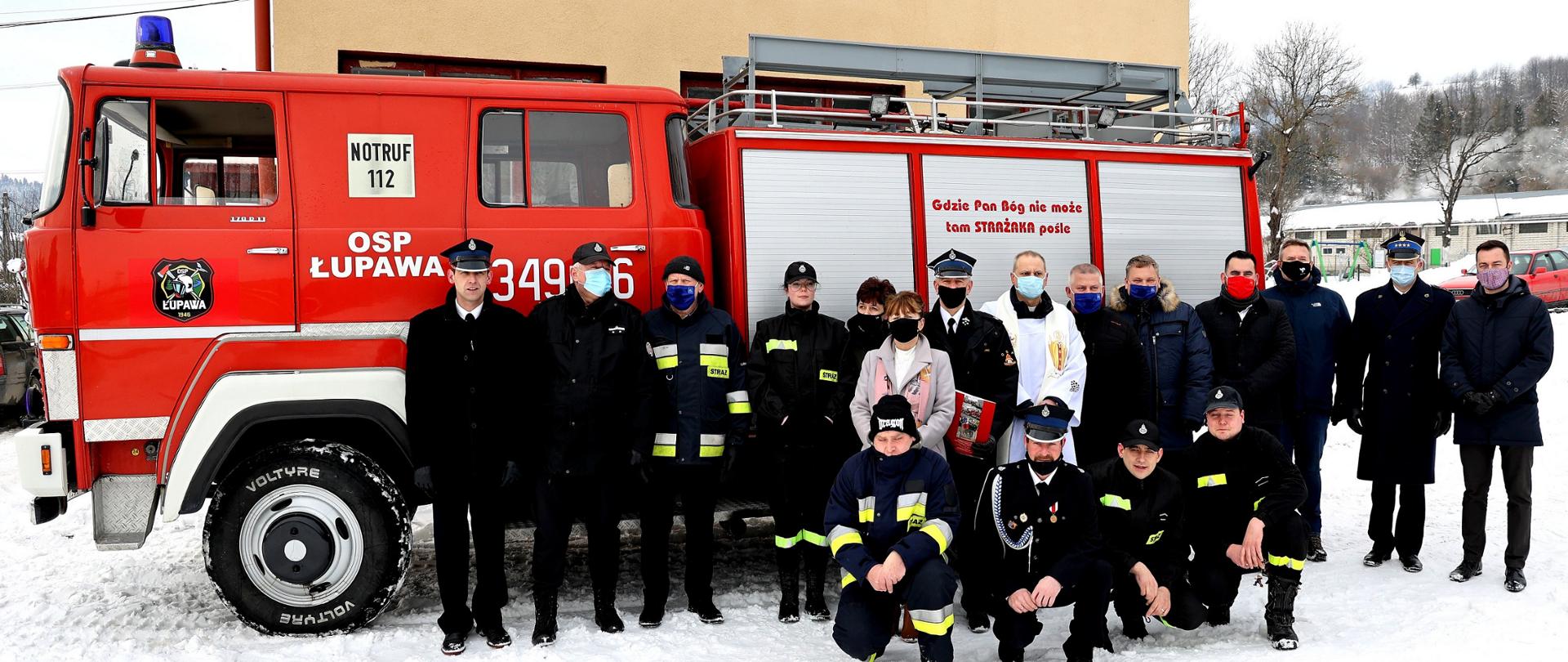 Zdjęcie grupowe na tle przekazanego samochodu pożarniczego. Przed samochodem stoją strażacy OSP ubrani w mundury galowe i koszarowe, Są tam również komendant powiatowy PSP, ksiądz oraz przedstawiciele lokalnych władz.