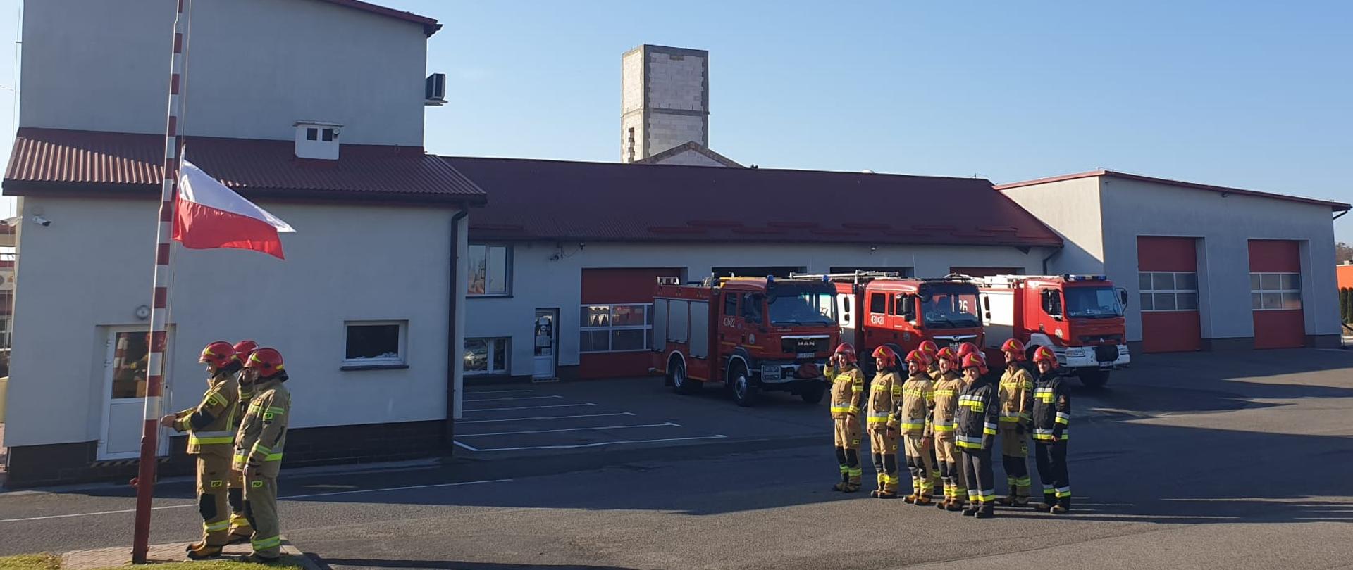 Na zdjęciu widać poczet flagowy wciągający flagę na maszt, a także zmianę służbową strażaków. Wszyscy strażacy ubrani są w umundurowanie specjalne, a na głowie mają hełmy. W tle widać budynek komendy oraz wozy strażackie
