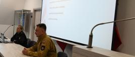 Za stołem siedzą funkcjonariusz w czarnym mundurze przed laptopem, obok strażak w mundurze w kolorze piaskowym. Obok baner Państwowej Straży Pożarnej.